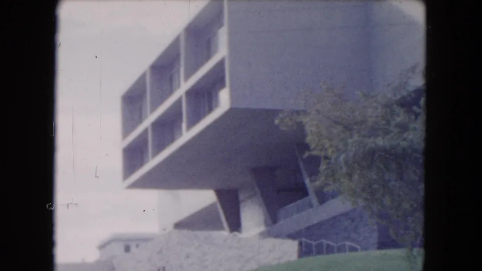 1961 South Carolina 8mm view of a white modern building with balcony-like openings, angled supports, a low wall, and trees at the base.