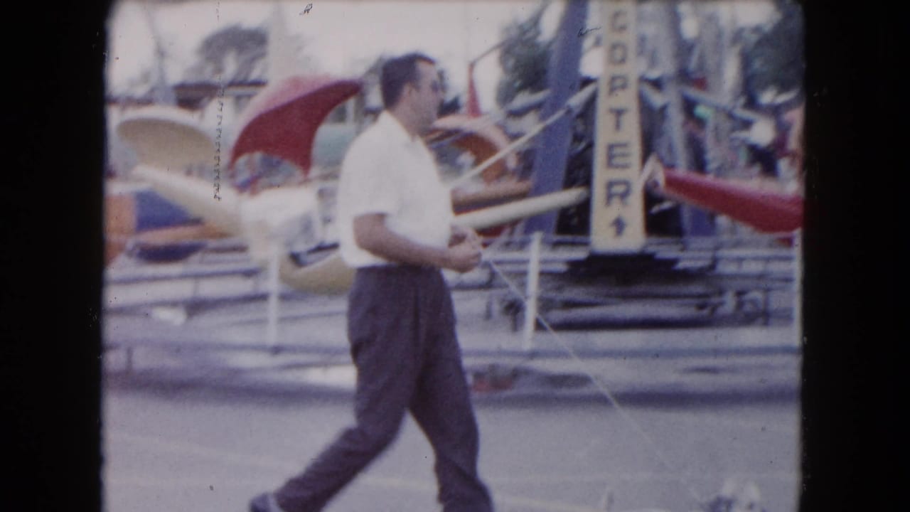 In 1961 South Carolina home-movie footage, a man in a white shirt walks through a small carnival beside helicopter-style ride cars.