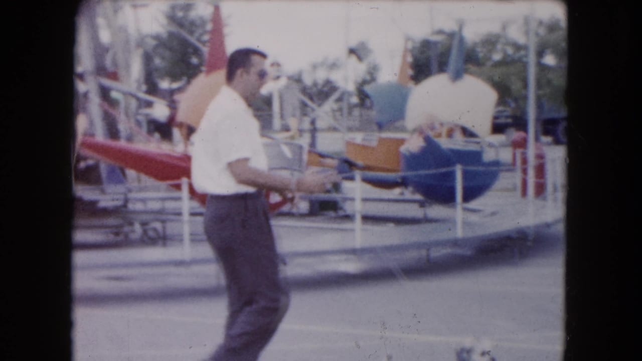 The man continues past railings and colorful ride cars at the small South Carolina carnival, with partial signage behind him.