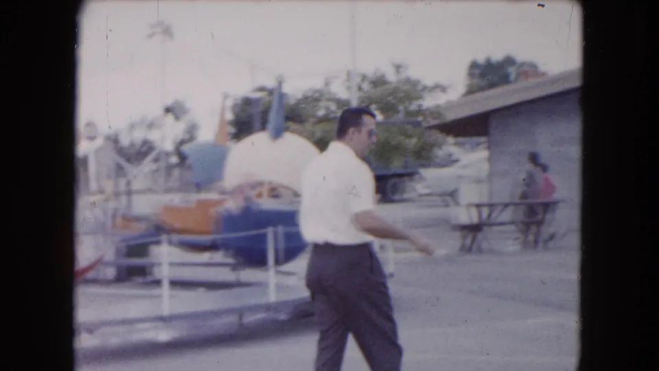 A handheld frame shows the man moving past carnival railings toward an open paved area with a bench and low building beyond.