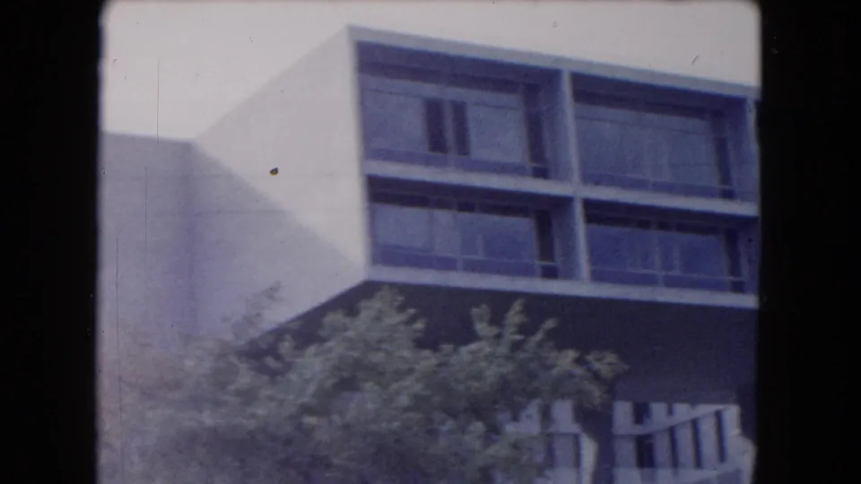 The clip shifts to a modern building facade with dark window bands, a projecting upper story, shrubs, and bright outdoor light.