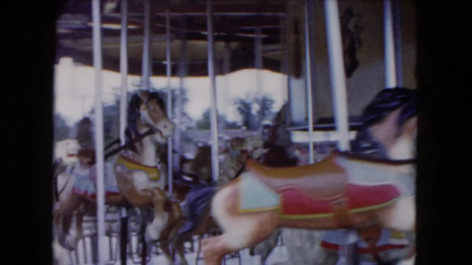 1961 South Carolina 8mm frame with a close painted carousel horse in front of other horses, poles, and partial riders.