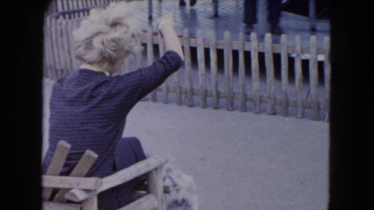 In 1961 South Carolina home-movie footage, a blonde woman in a dark dress sits outdoors and gestures toward a small dog by a fence.