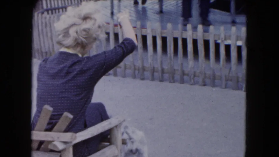 In 1961 South Carolina home-movie footage, a blonde woman in a dark dress sits outdoors and gestures toward a small dog by a fence.
