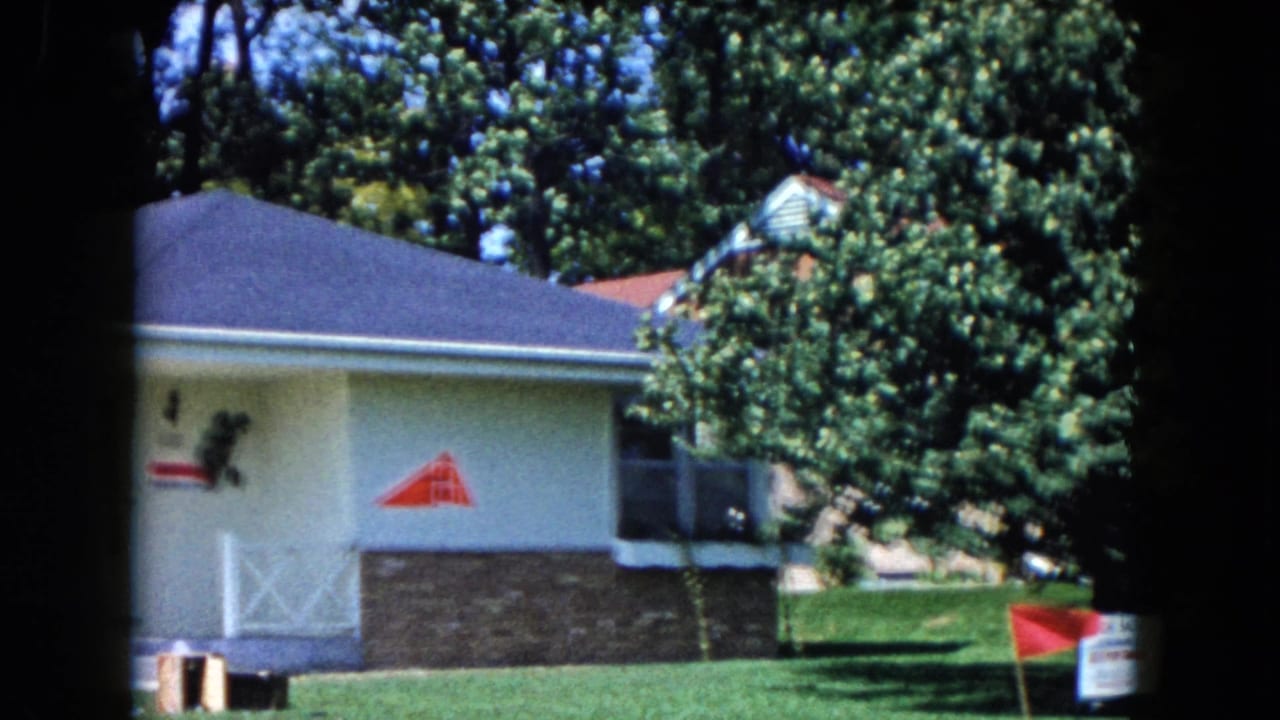 1961 South Carolina home-movie frame showing a white suburban ranch house, grass lawn, trees, red marker, and partial real-estate signage.