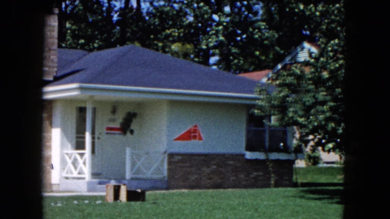Light-colored single-story house with porch, brick foundation, lawn, trees, and a red wall logo.