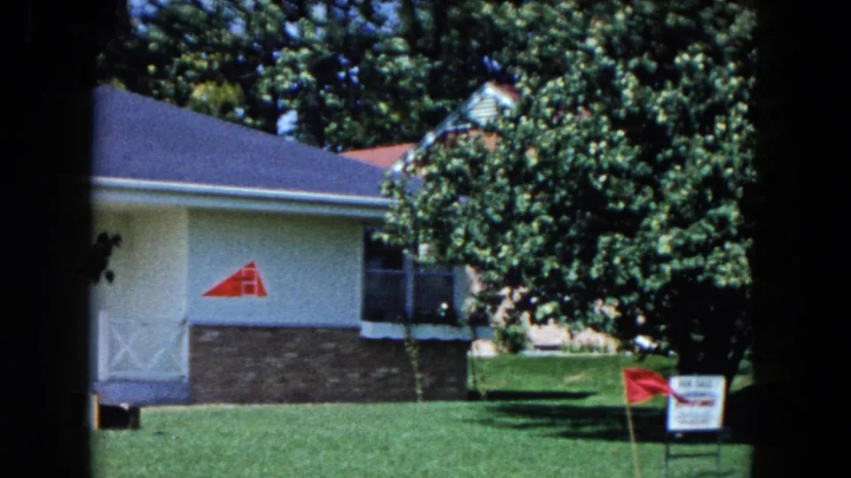 1961 South Carolina frame showing the side of a neat house, lawn, trees, a red wall logo, and a small real estate sign with a red flag.