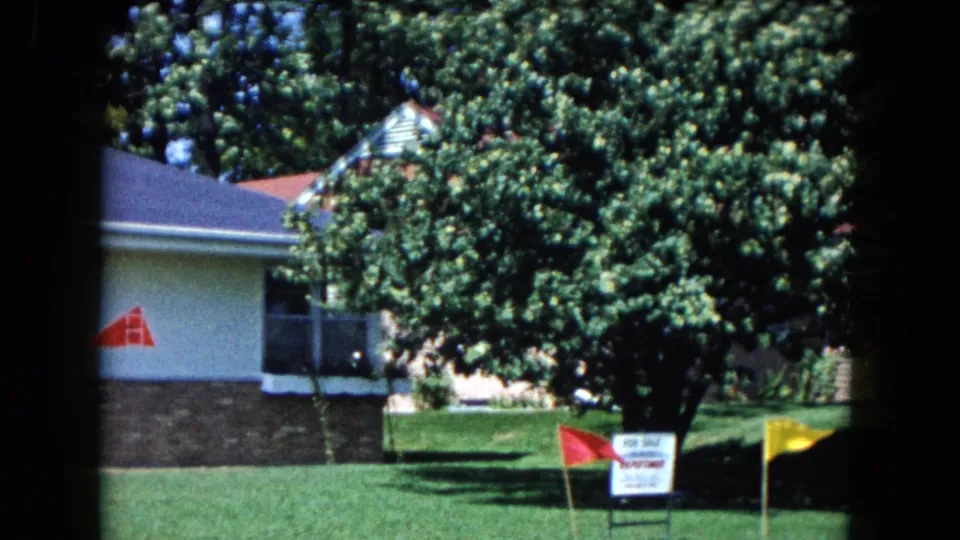 1961 South Carolina home-movie frame with the house partly obscured by trees, a manicured lawn, real estate sign, and bright yard flags.