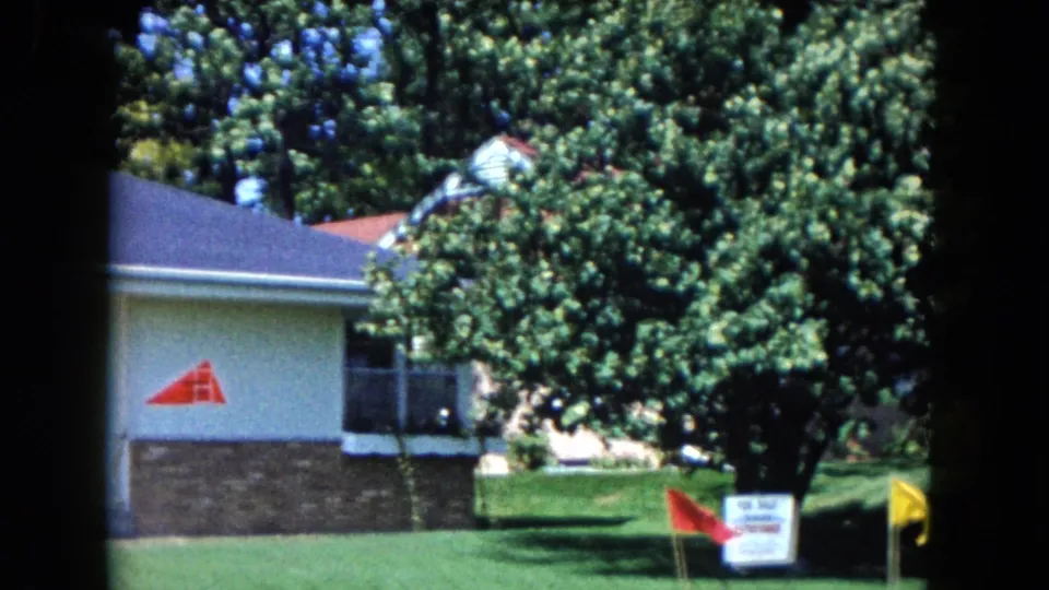 1961 South Carolina frame showing a residential lawn with a real estate sign, red and yellow flags, mature tree cover, and part of the house.
