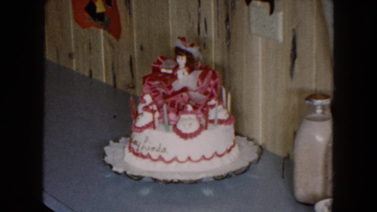 1961 South Carolina home-movie frame showing a white birthday cake with pink trim, doll topper, and milk bottle on a blue countertop.