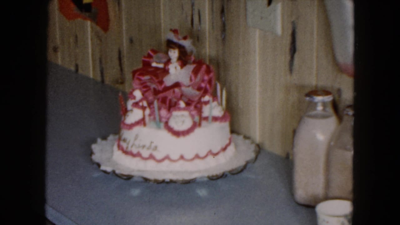 Close 8mm view of Linda's decorated birthday cake with pink ornamentation, doll topper, and a milk bottle in a private interior.