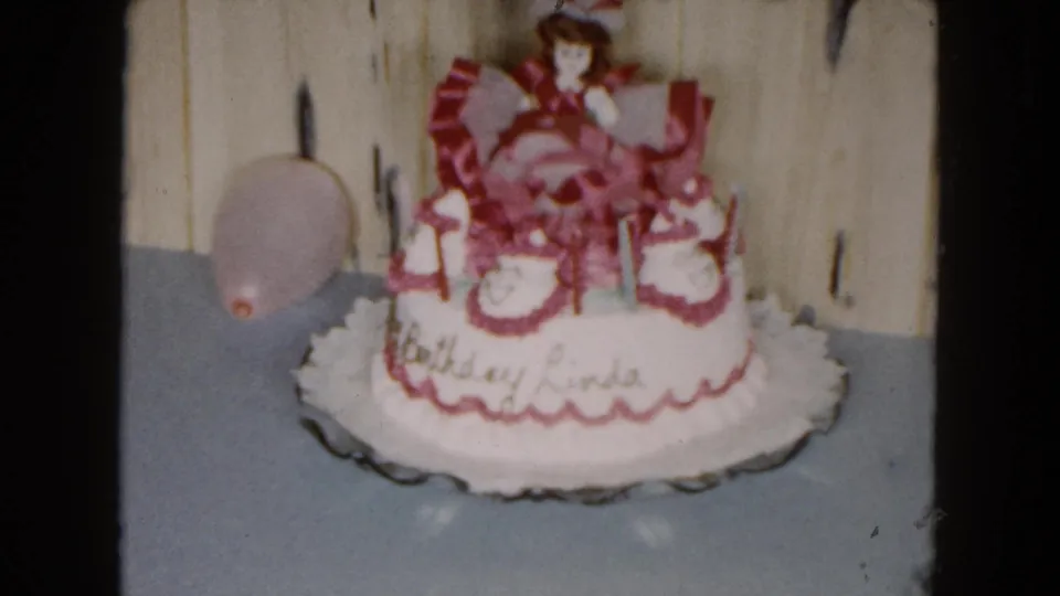 Closer frame of the 1961 birthday cake showing pink trim, a doll topper, cursive writing for Linda, and a pink balloon at the side.