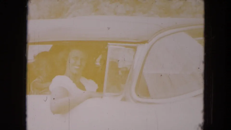 A woman smiles from inside a car during a 1961 South Carolina family vacation home movie, seen through faded 8mm film exposure.