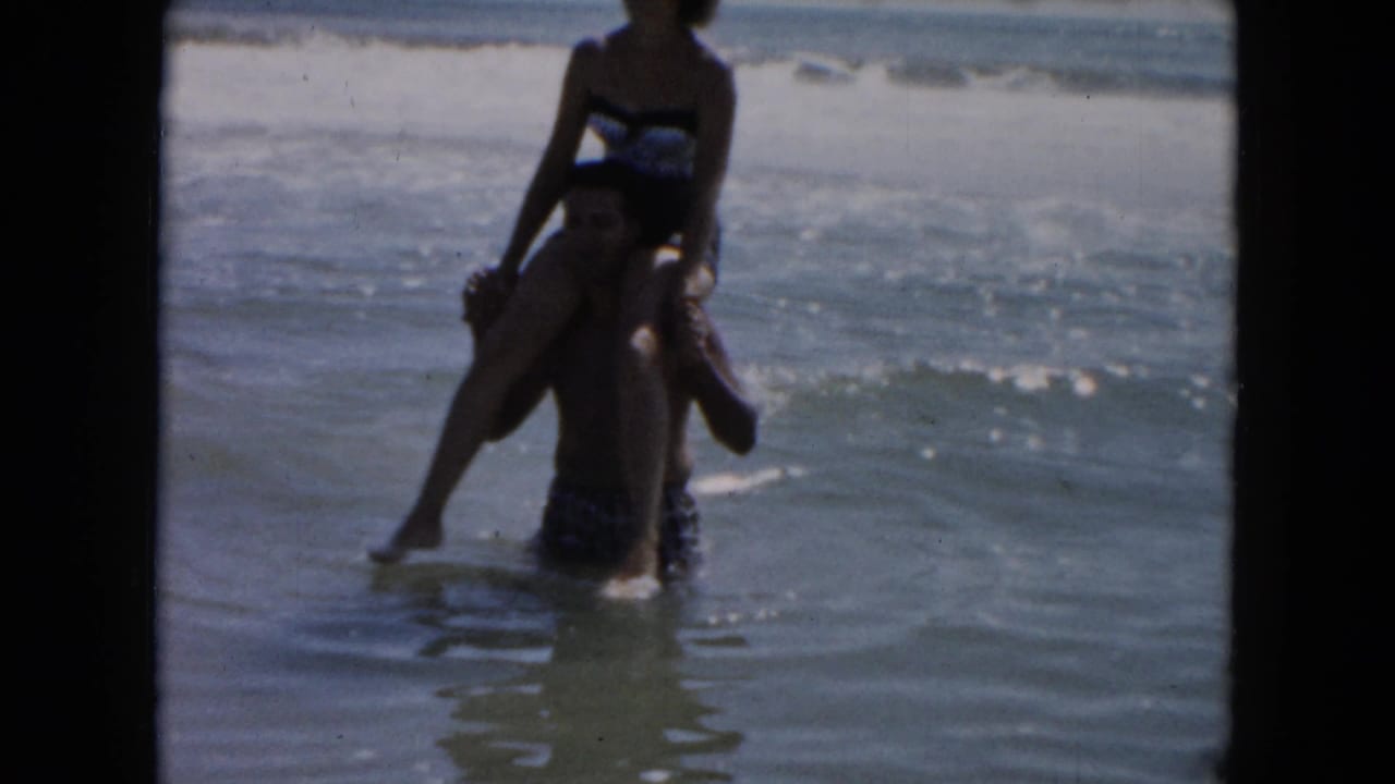 Man carries a woman on his shoulders in shallow ocean water.