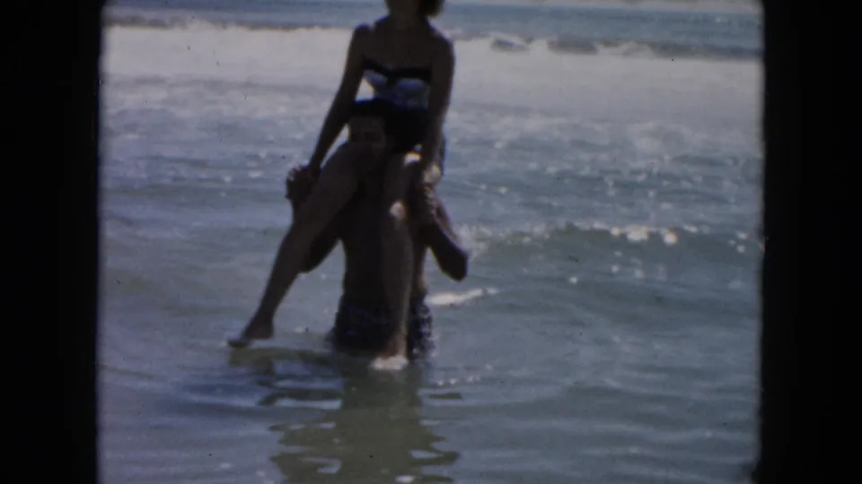 In 1961 South Carolina beach footage, a man wades in the surf while carrying a woman on his shoulders.
