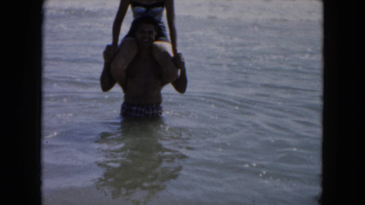 A man in swim trunks carries a woman on his shoulders in shallow South Carolina surf in 1961 home-movie footage.