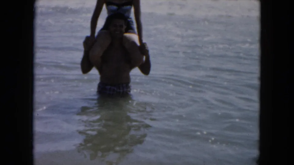A man in swim trunks carries a woman on his shoulders in shallow South Carolina surf in 1961 home-movie footage.