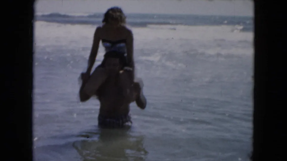The woman sits astride the man's shoulders while he stands in the shallow ocean, with soft 8mm grain and faded color.