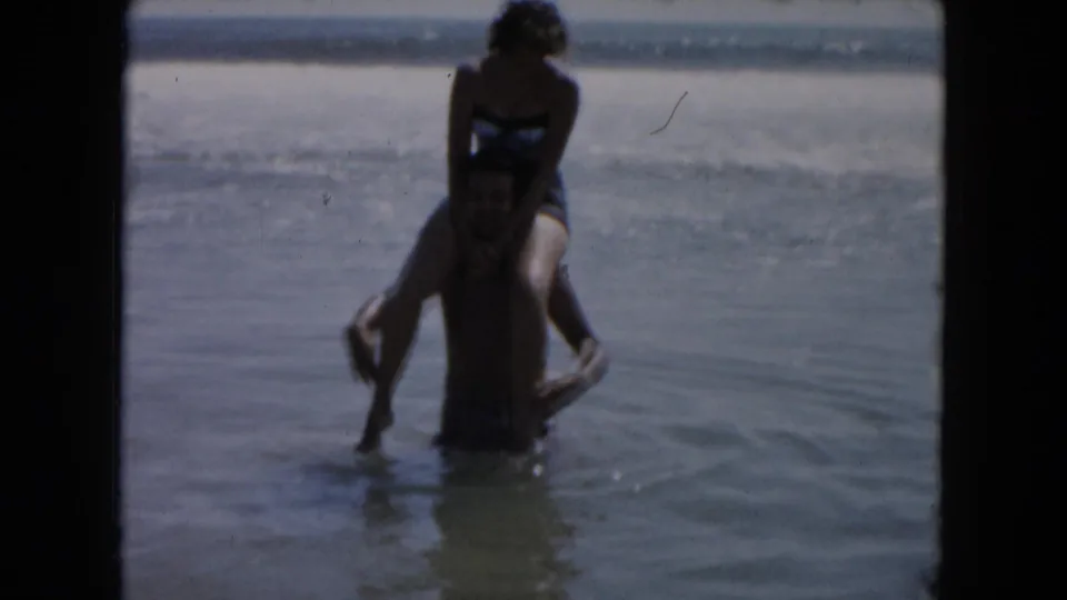 In 1961 South Carolina beach footage, a man stands in shallow surf with a woman seated on his shoulders.
