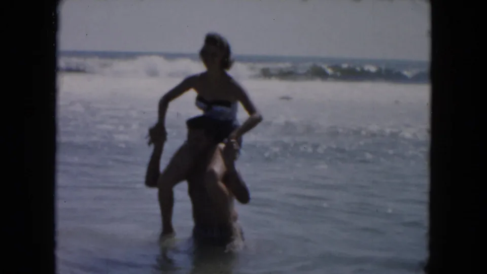 A woman in a dark swimsuit balances on a man's shoulders in shallow South Carolina surf, supported by 1961 metadata.