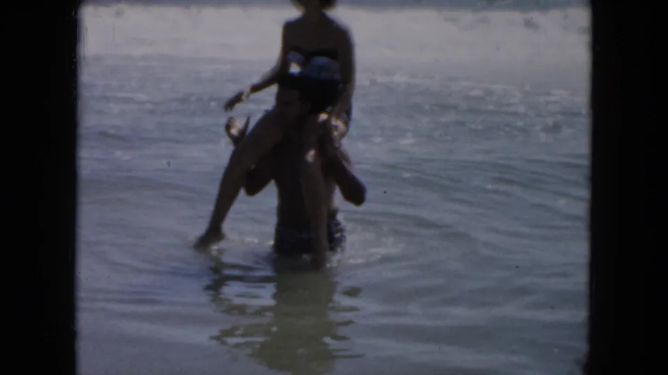 The home-movie frame shows the couple in shallow surf, with the woman on the man's shoulders and the ocean stretching behind them.