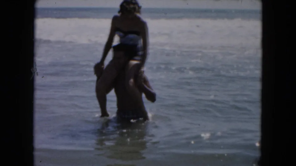 A 1961 South Carolina beach home-movie frame shows a man in the surf carrying a woman on his shoulders near low waves.