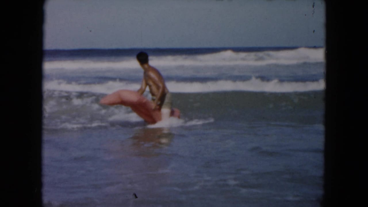 Young man in swim trunks appears in the 1961 South Carolina surf, holding or mounting a light pink inflatable float near breaking waves.