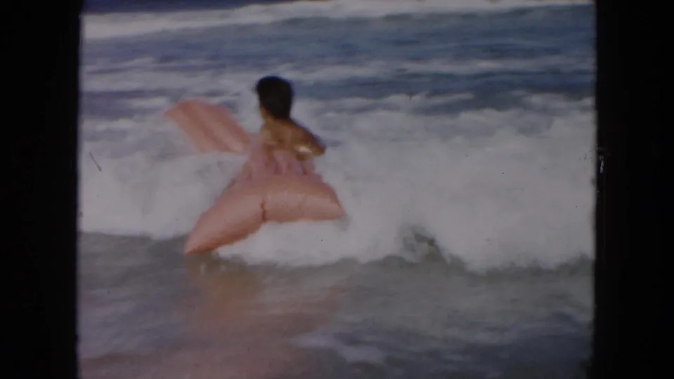 The man is surrounded by white surf while the light pink inflatable float tilts upward in the South Carolina ocean scene.