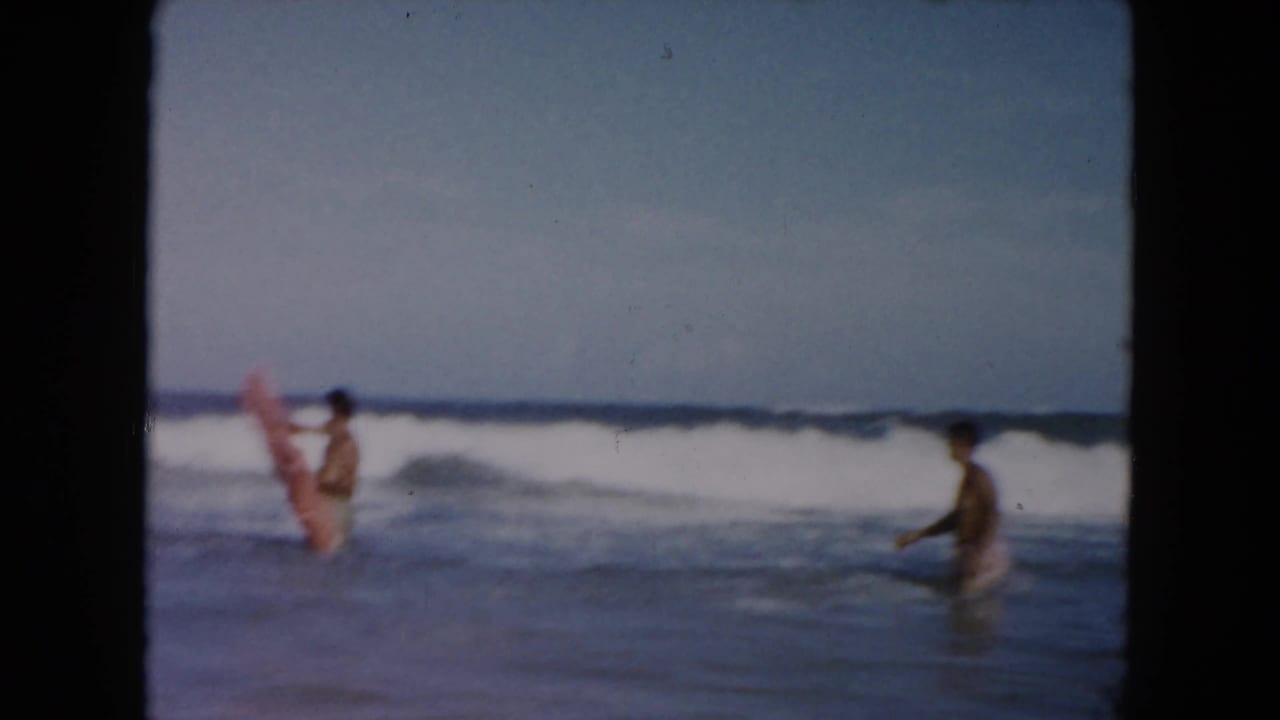 Two young men in ocean surf with a pink inflatable raft near breaking waves.