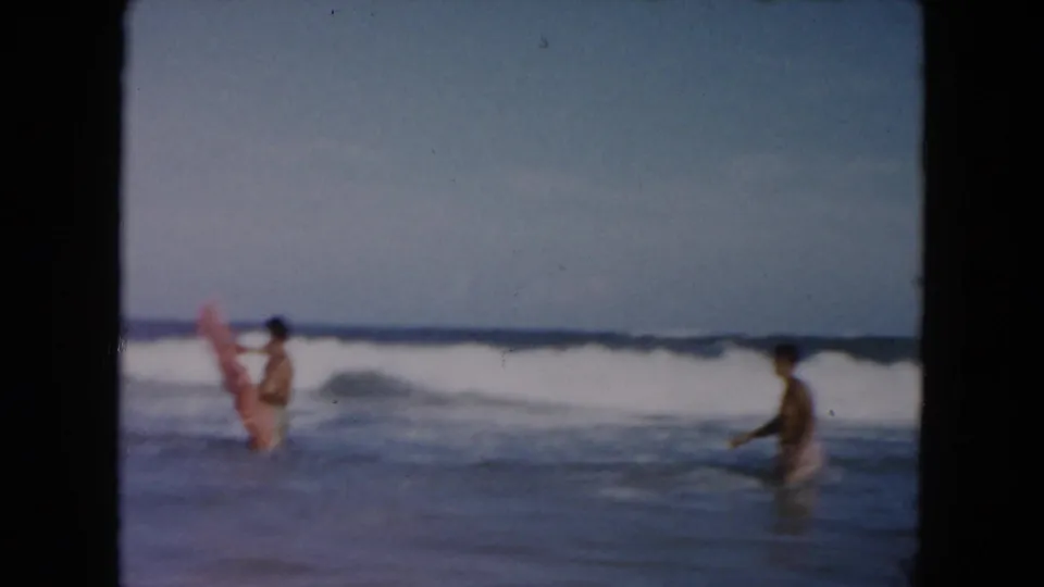 1961 South Carolina beach home-movie frame showing two young men in the surf, one holding a pink inflatable raft as waves break behind them.