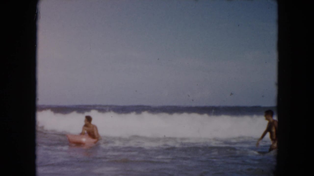 1961 South Carolina beach footage frame with a young man on a pink inflatable raft and another person standing at the edge of the breaking surf.