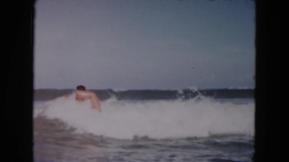 1961 South Carolina beach frame showing a young man and pink inflatable raft partly hidden by a breaking wave in soft 8mm color.