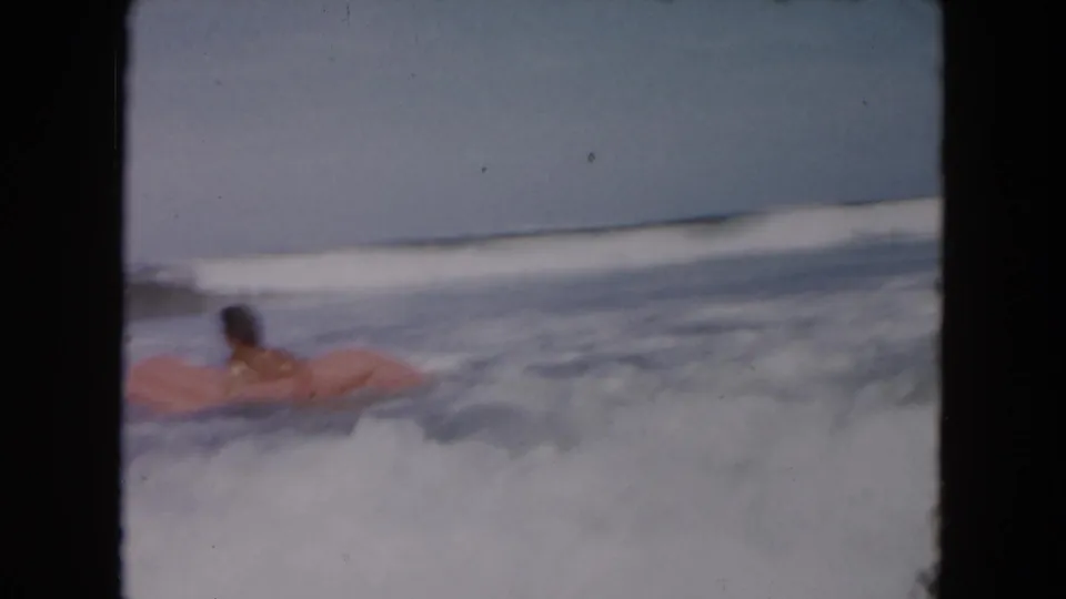 1961 South Carolina beach home-movie frame showing a young man prone on a pink inflatable raft as surf washes around him.