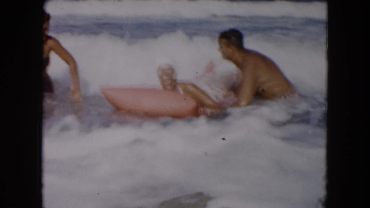 Adults in ocean surf play around a pink inflatable raft as waves break nearby.