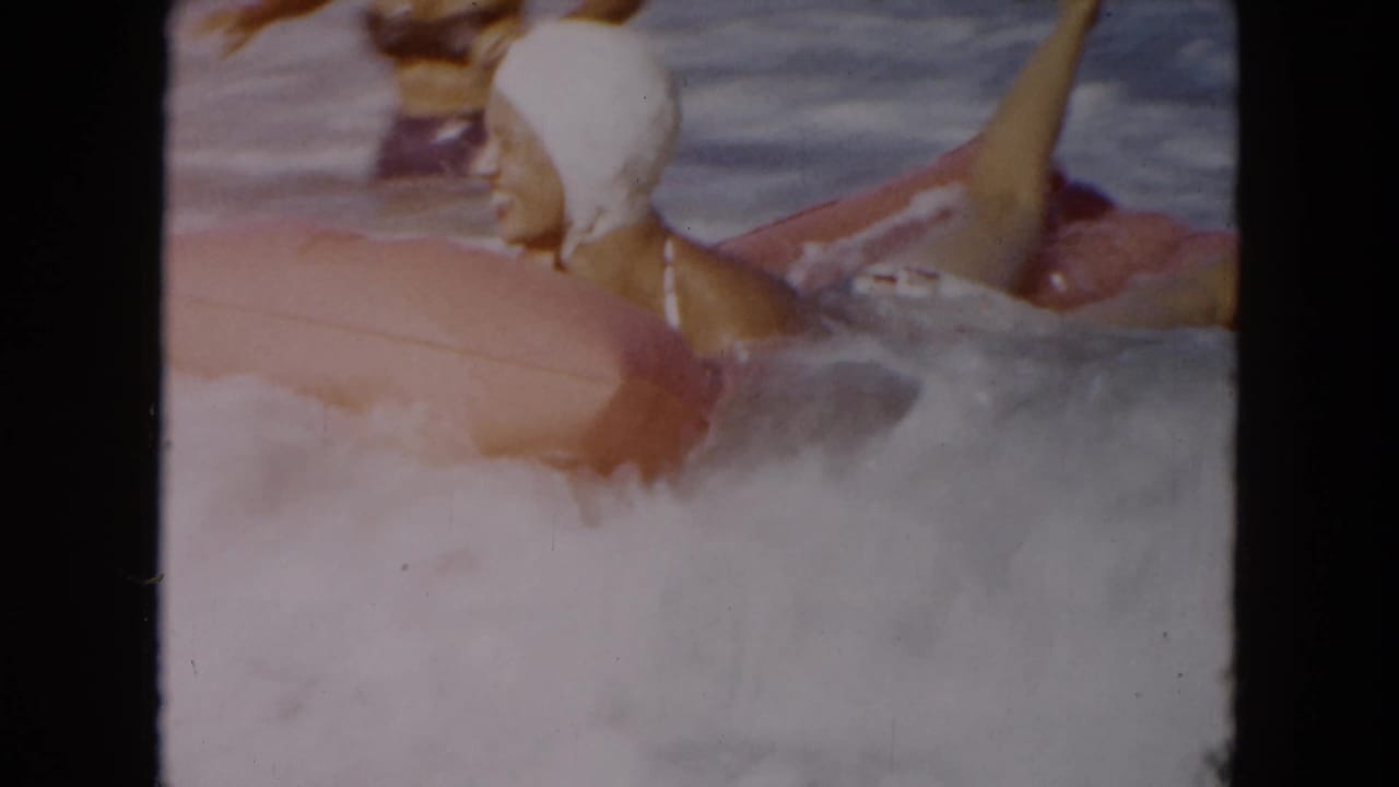 Woman in a white swim cap rides through foamy surf beside a pink inflatable raft.