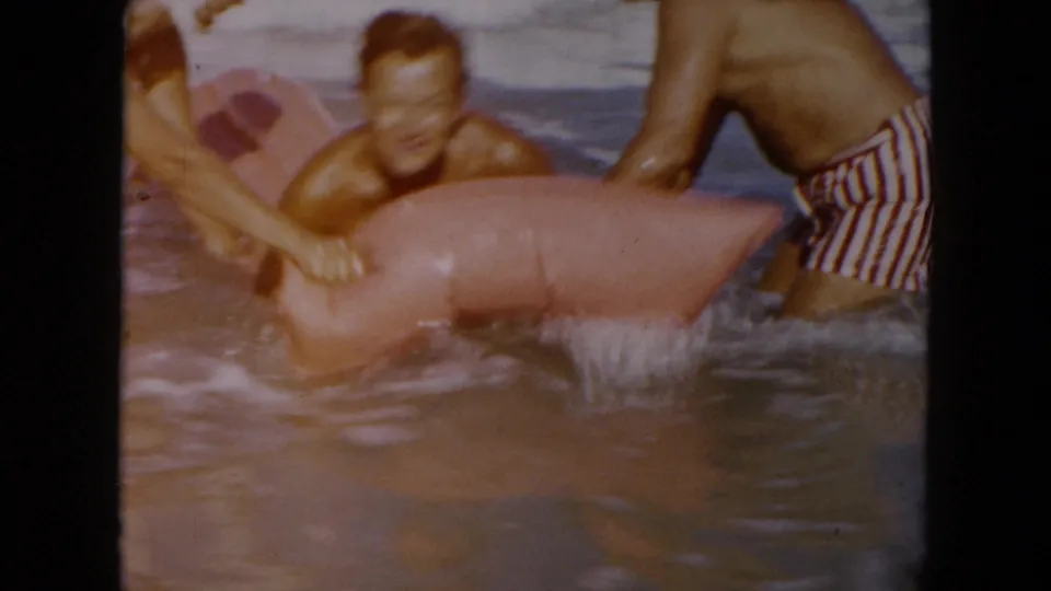 Adults steady a pink inflatable raft in shallow South Carolina surf, with striped swim trunks visible in the 1961 8mm frame.