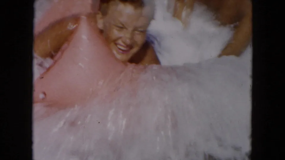 A smiling woman holds onto a pink inflatable raft as South Carolina surf splashes around her in 1961 home-movie footage.