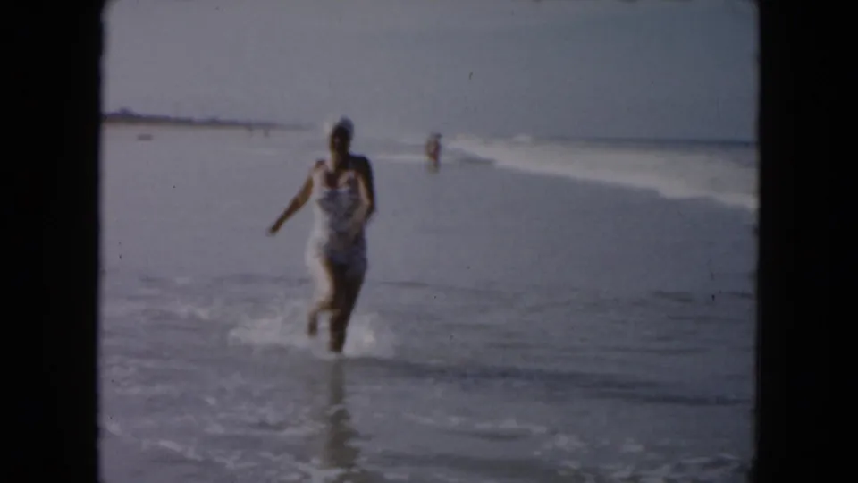 In this 1961 South Carolina beach frame, a woman in a patterned one-piece swimsuit runs through shallow waves near the shoreline.