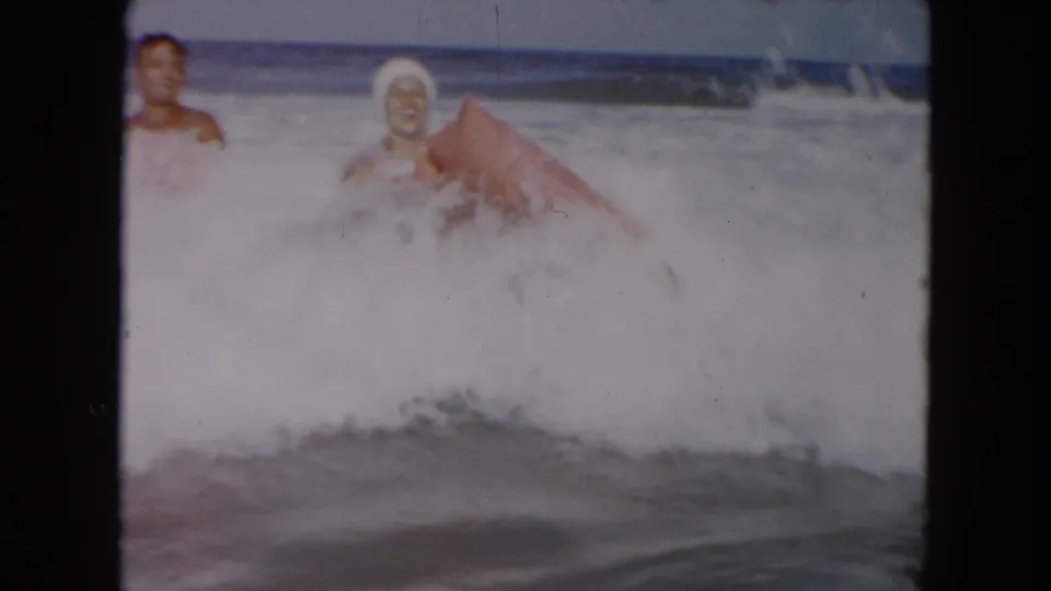 Two people appear in churning surf at the beach, with a red float or board partly visible among the waves.