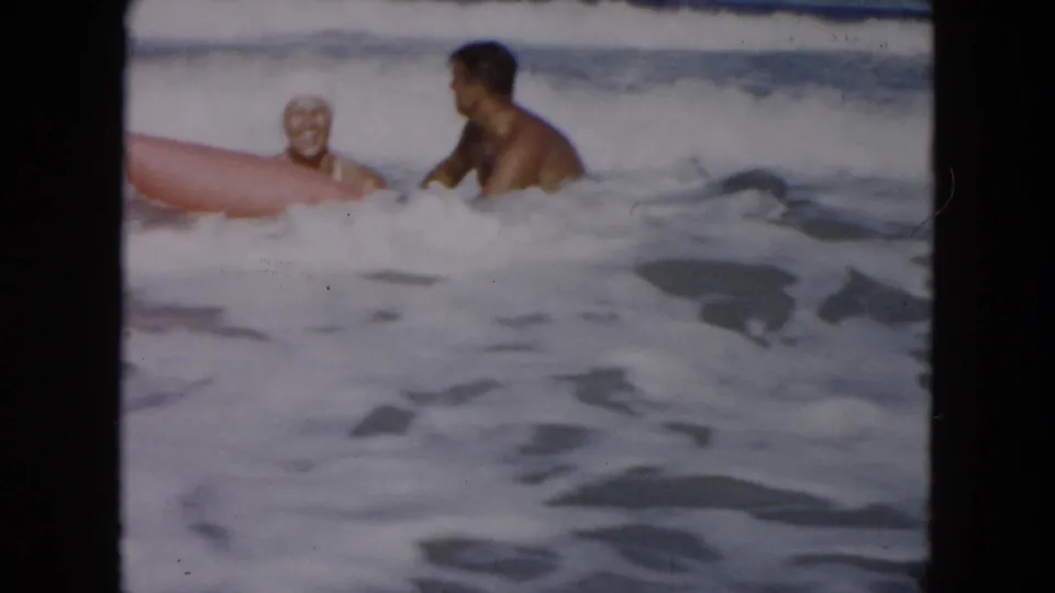 Two adults in the surf face each other beside a red float or board as waves break behind them at the beach.