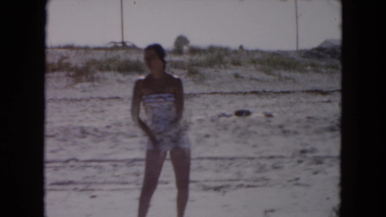 1961 South Carolina home-movie frame of a woman in a patterned one-piece swimsuit standing on a sandy beach near low dunes.