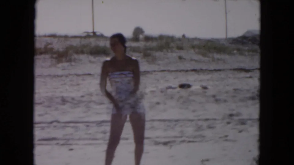 1961 South Carolina home-movie frame of a woman in a patterned one-piece swimsuit standing on a sandy beach near low dunes.