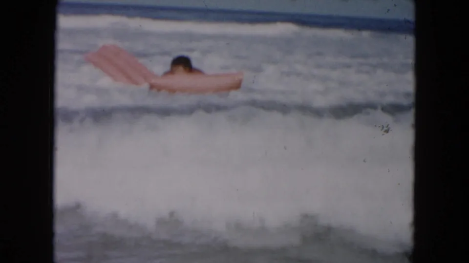 1961 South Carolina home-movie frame of a swimmer on a pink inflatable float as small waves break around them.