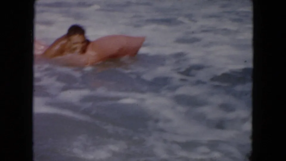 1961 South Carolina beach frame showing a swimmer leaning on a pink inflatable float in moving ocean water.