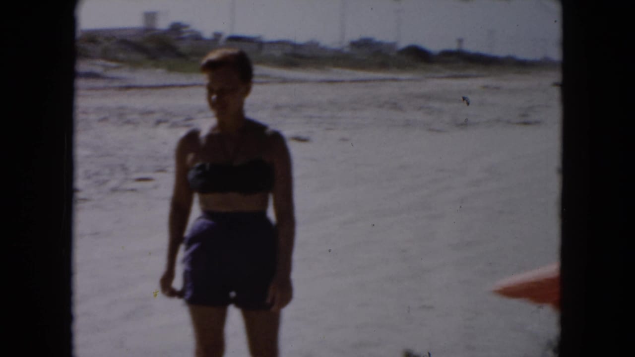 1961 South Carolina 8mm frame showing a woman in a dark two-piece swimsuit standing on open sand with water and low dunes behind her.