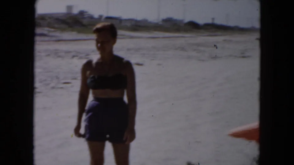 1961 South Carolina 8mm frame showing a woman in a dark two-piece swimsuit standing on open sand with water and low dunes behind her.