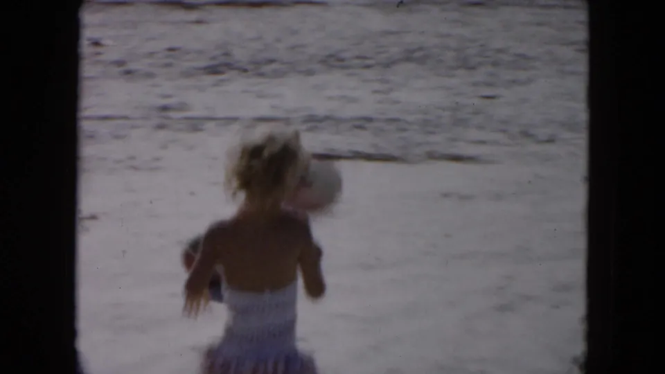 1961 South Carolina beach frame showing a person in a patterned swimsuit from behind with a ball on the sand nearby.