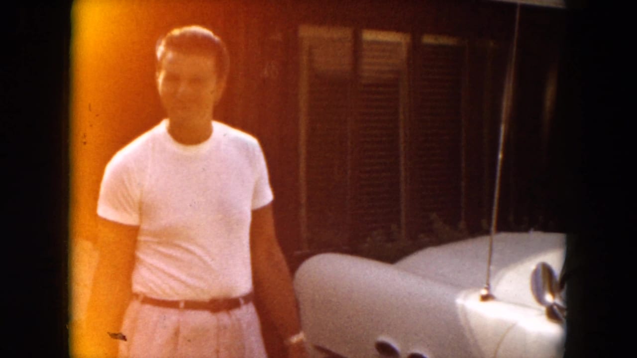 Man in a white T-shirt stands beside a light-colored classic car.