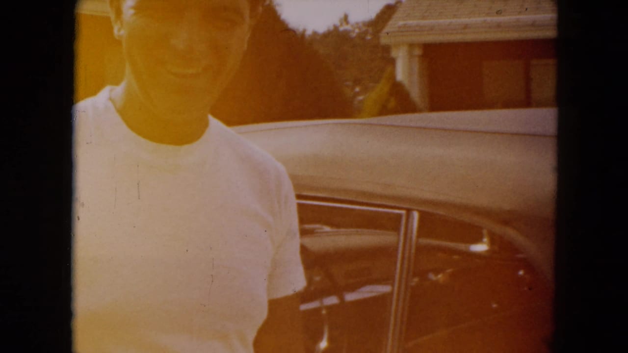 A smiling man in a white T-shirt stands near the passenger side of a classic car in the South Carolina home-movie scene.