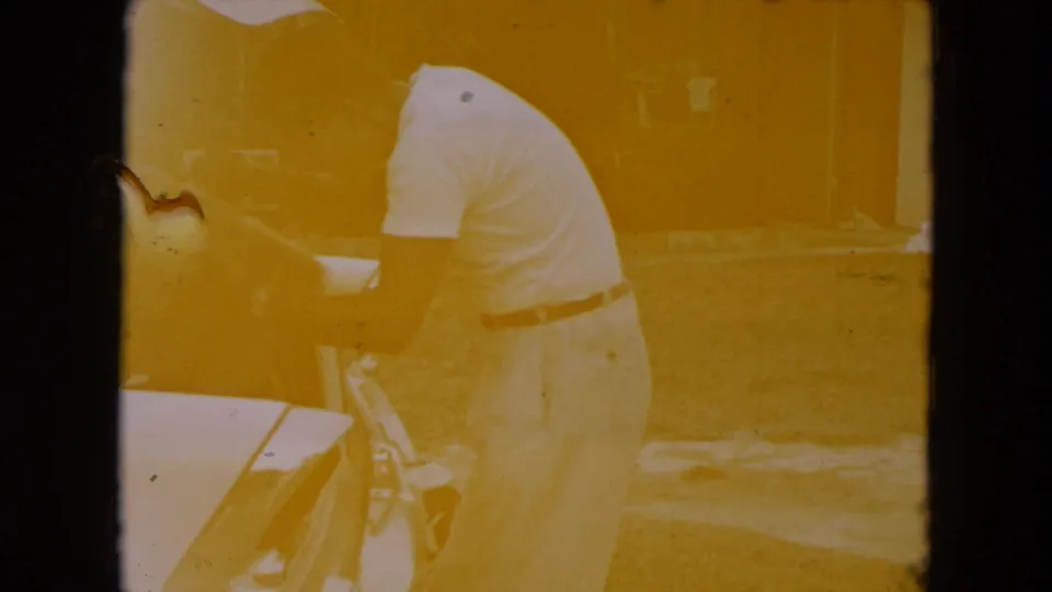 The man bends at the rear of the classic car, appearing to place luggage into the open trunk during the 1961 travel scene.